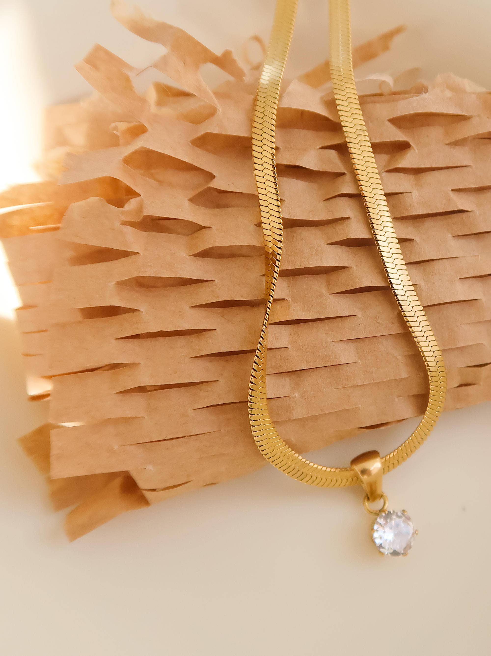 Flat lay: Close-up of 18K gold snake chain necklace with cubic zirconia round pendant on white surface